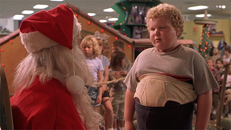 Chubby kid with curly hair and his shirt over his belly talking to Santa