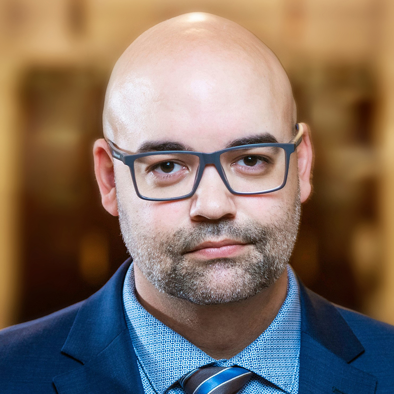 White bald man with glasses wearing a blue suit, shirt and tie.