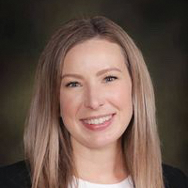 Portrait of a smiling white women with blonde hair in a black blazer and white blouse in front of a dark background.
