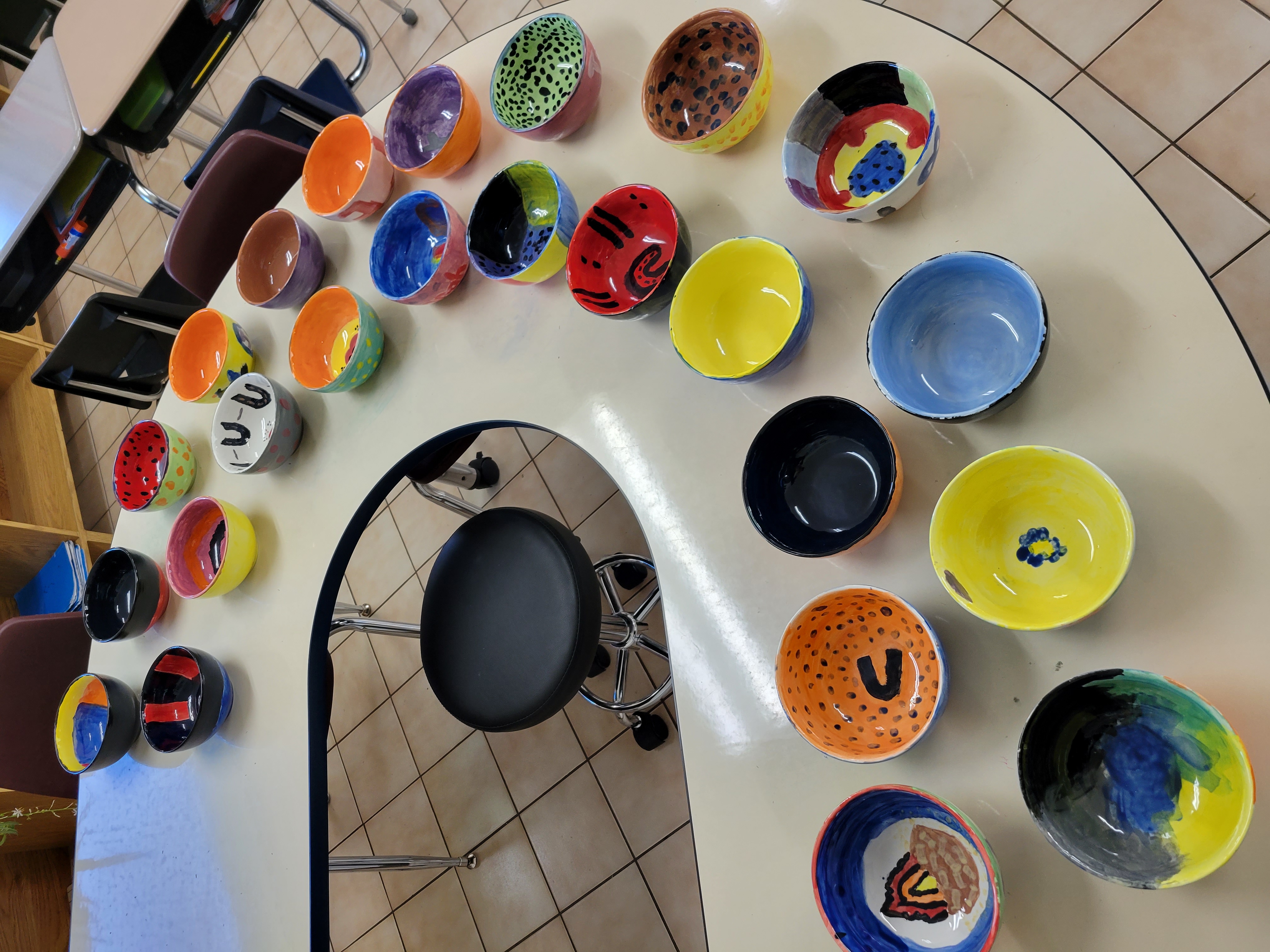 A round desk with a stool in the middle. On the desk are 26 multicolour bowls, student pottery projects.