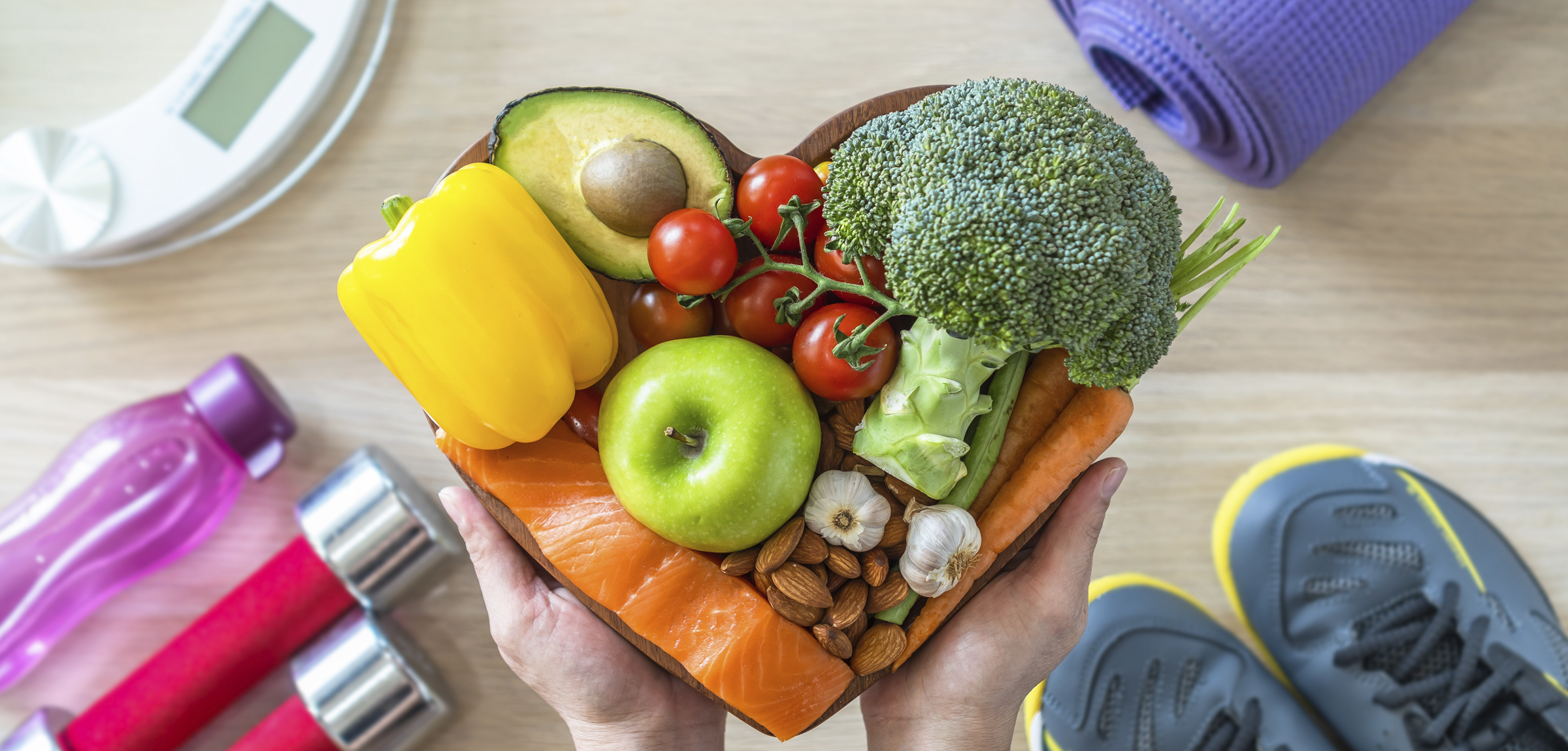 The background is a light wood table or floor. There is a scale, a yoga mat, weights, and running shoes in each corner. In the foreground two hands hold fruits, vegetables, nuts and salmon. These items of food are arranged in a heart shape. 