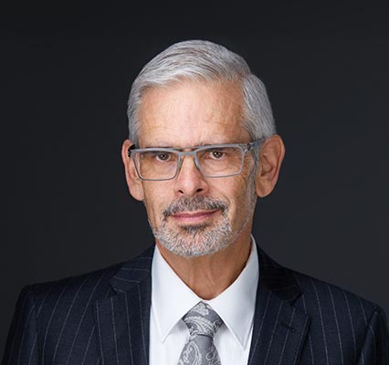 Grey haired man in silver glasses wears a dark suit infront of black background