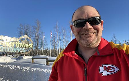 Man wearing a red coat poses in front of Northwest Territories sign on a clear winter day