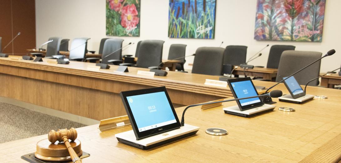 Provincial Executive Council chamber a large round wooden desk lined with microphones and chairs facing the center and paintings of nature on the wall.