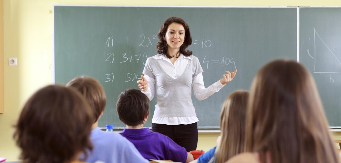 White female with brown hair standing in front of a chalkboard teaching students in the foreground